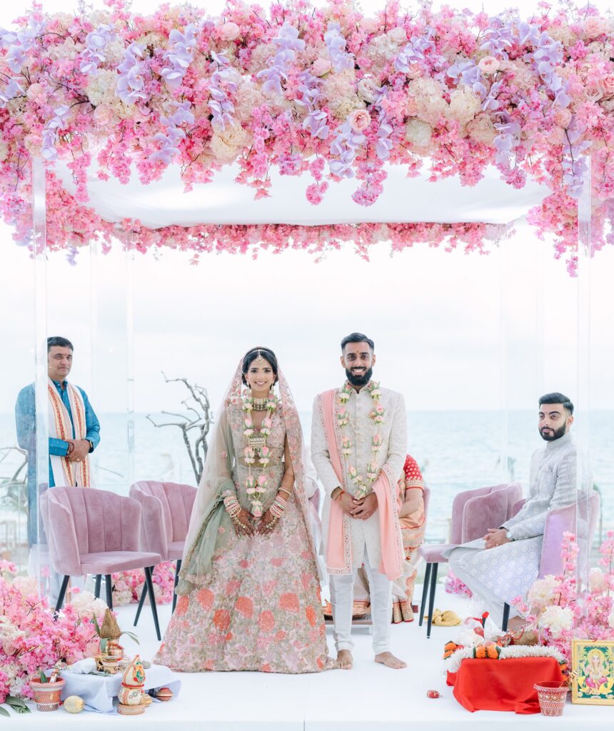 Bride and groom with garlands standing under a pink floral mandap during their Hindu wedding ceremony in Greece