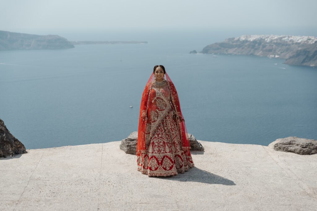South Asian bride in a red and gold lehenga on a clifftop overlooking the Santorini caldera in Greece