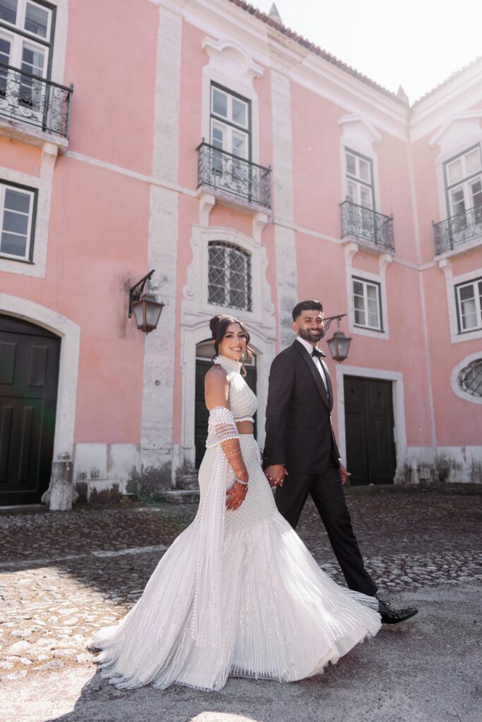 South Asian bride and groom walking past a pink palace façade in Portugal
