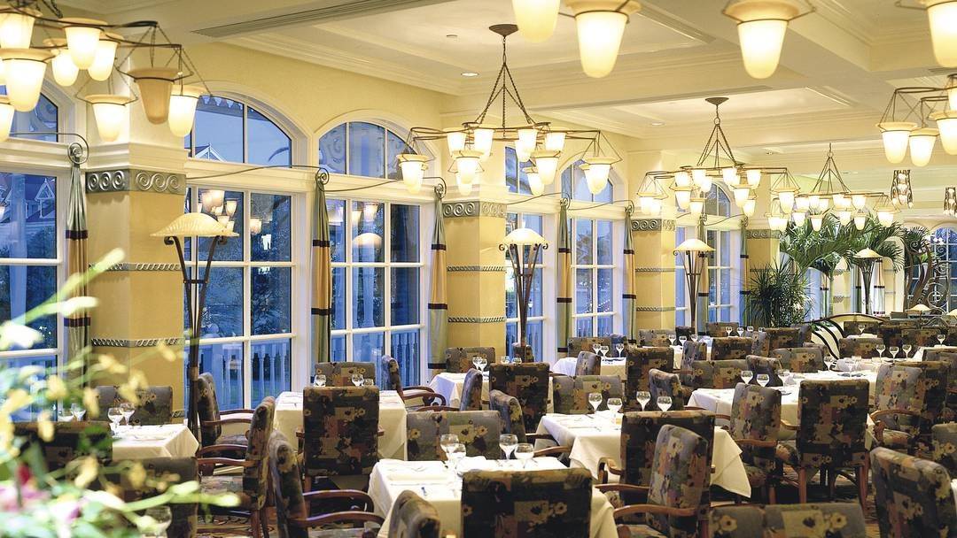 A well-lit restaurant interior with large windows, floral patterned chairs, and tables set with white tablecloths and glassware. Multiple chandeliers hang from the ceiling.